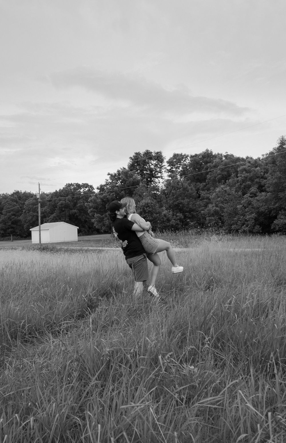Two people playfully interact in a black and white photograph taken in a rural field with trees in background.