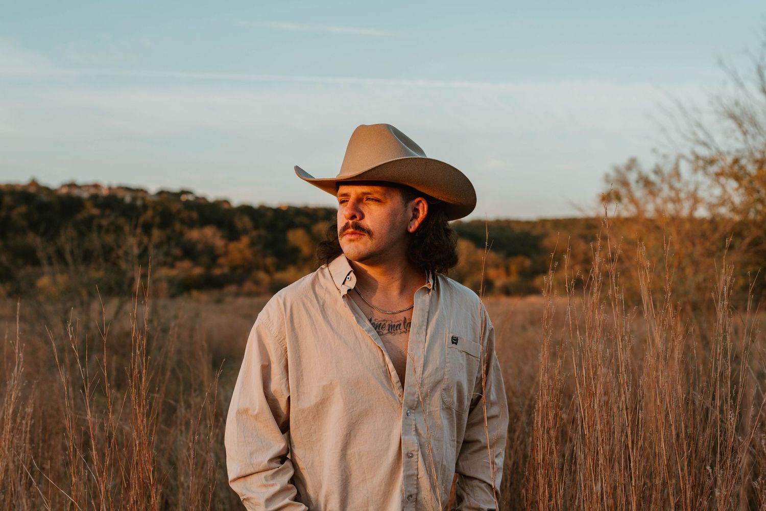 A person wearing a cowboy hat and light colored shirt stands in a field of tall grass at sunset.