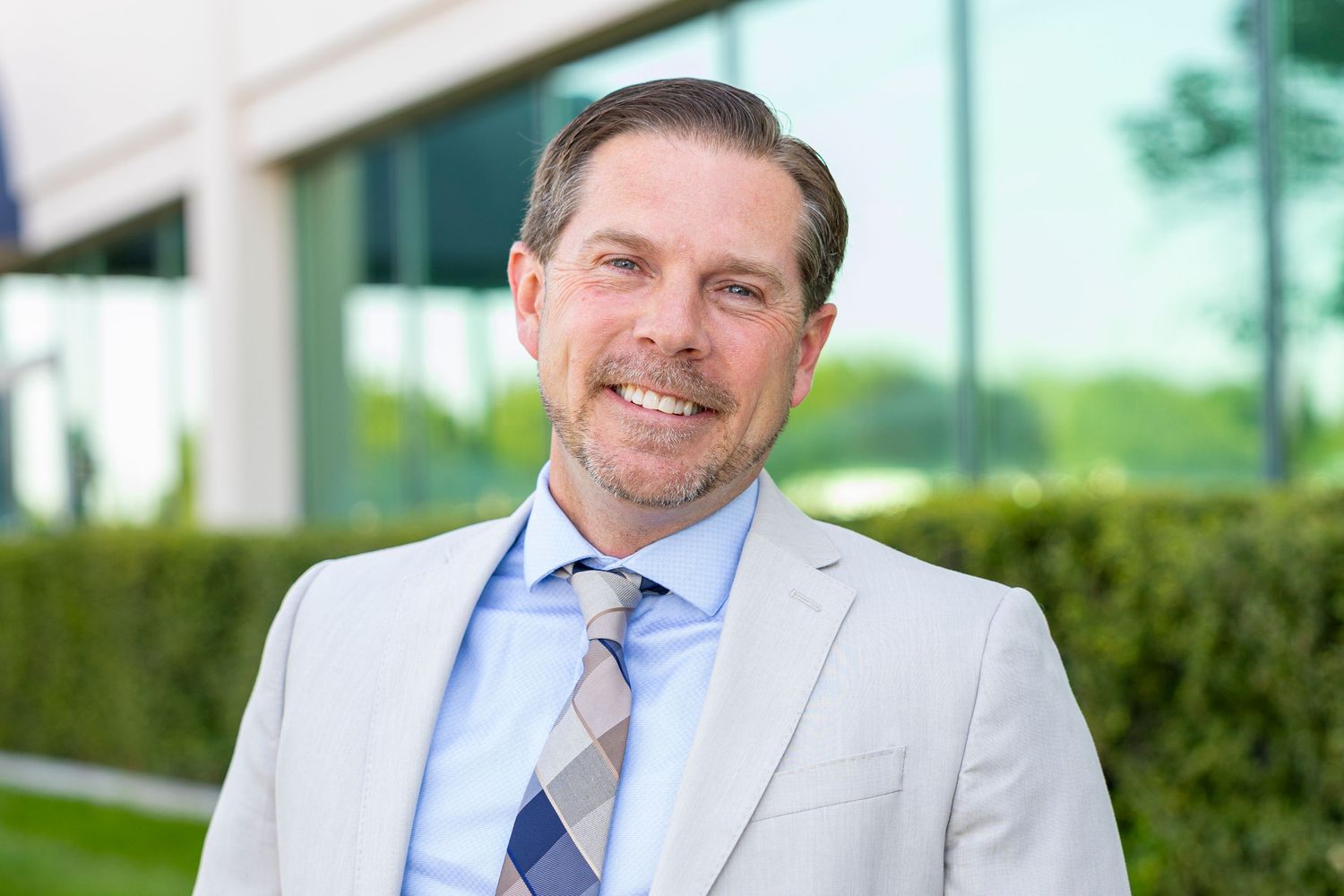Professional wearing a light gray suit and blue tie smiles while standing outdoors near greenery.