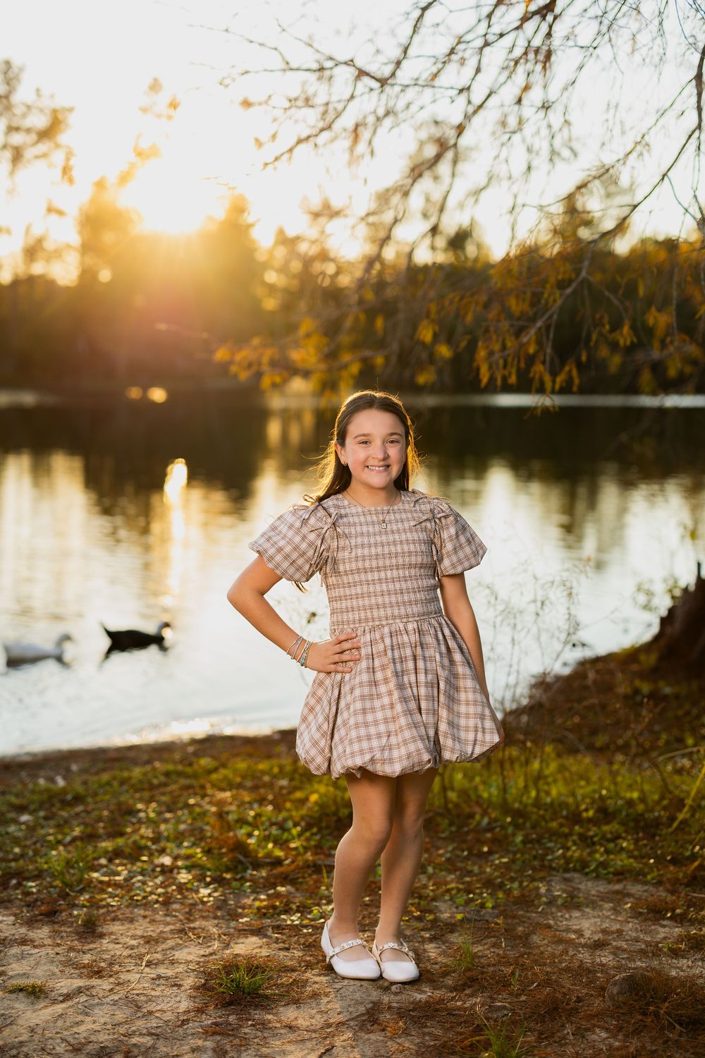 Young girl in checkered dress poses by lake during golden hour autumn photoshoot.