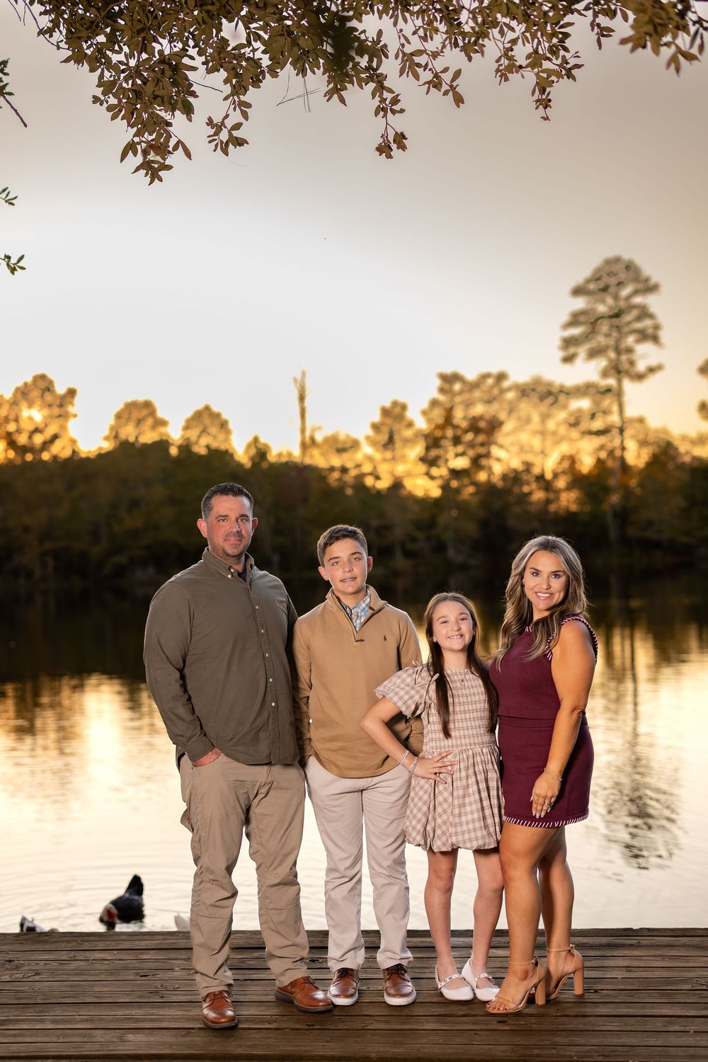Family sits together on wooden dock during sunset with lake and trees in background.
