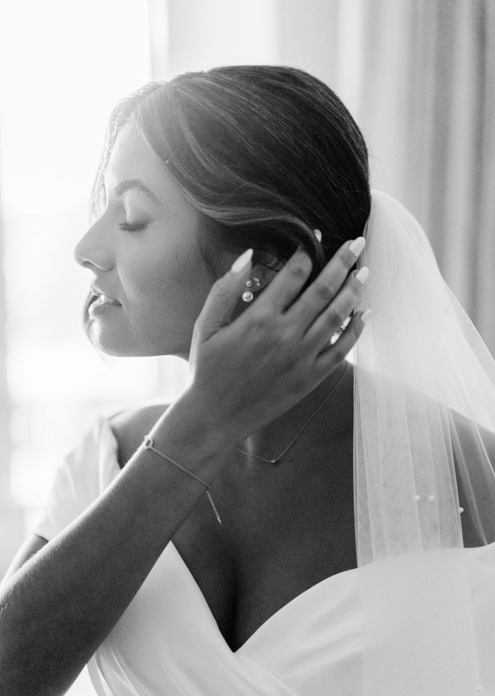 Bride wearing delicate jewelry and veil in soft window light during wedding preparations.