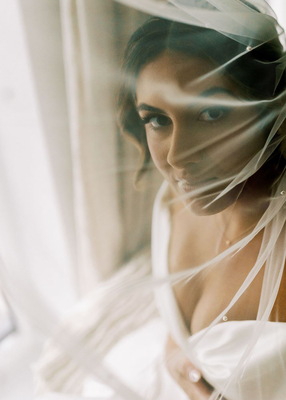 Ethereal bridal portrait through sheer white veil in natural window light.