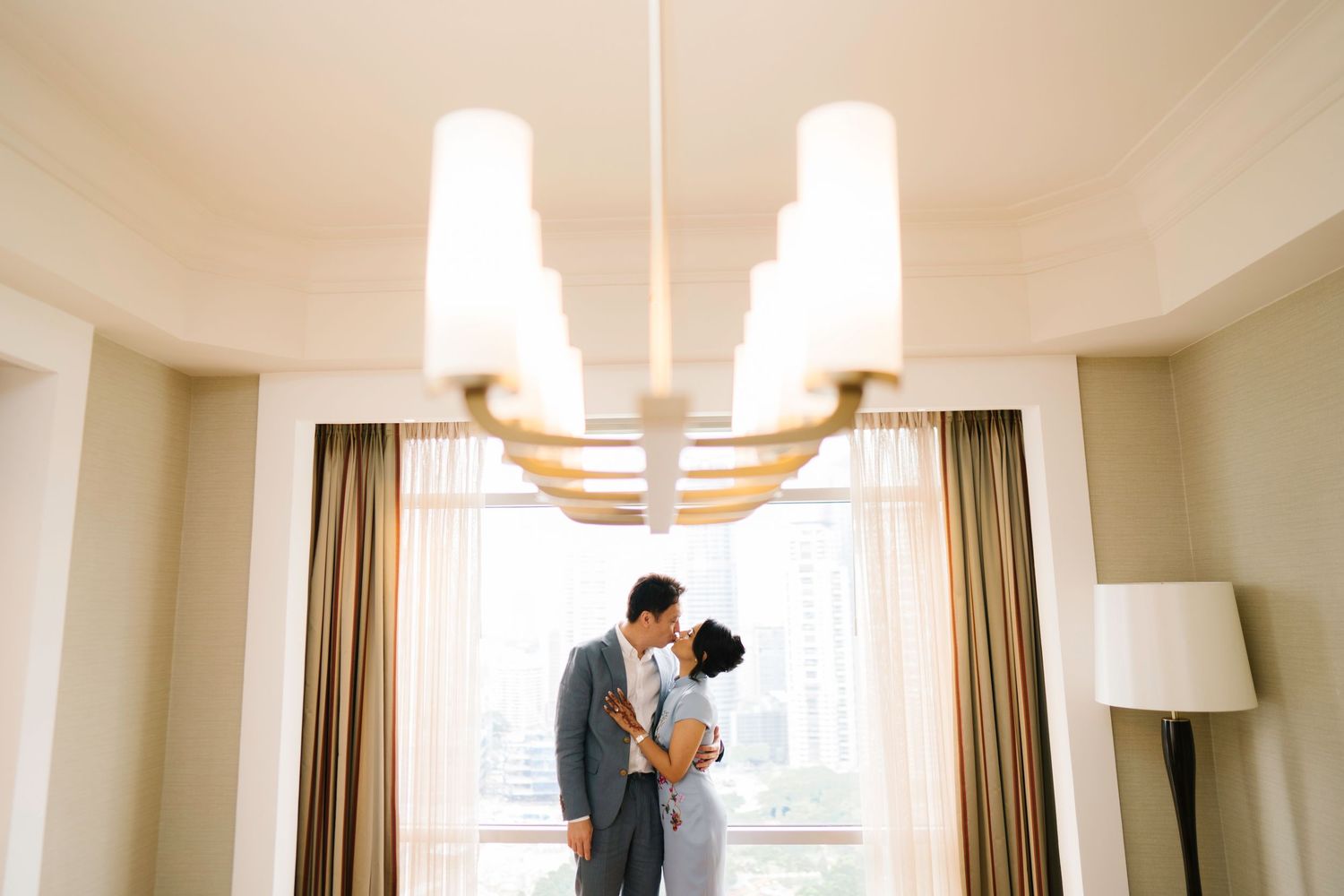 Couple shares romantic kiss under elegant chandelier by bright hotel room window.