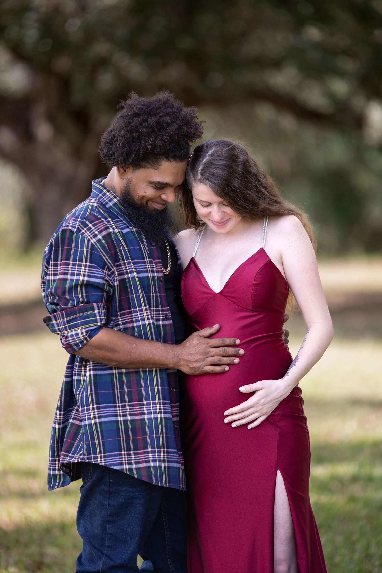 A couple in a plaid shirt and burgundy dress share intimate moments during an outdoor maternity photoshoot.