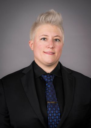 Professional headshot portrait in black suit and blue patterned tie against gray background.