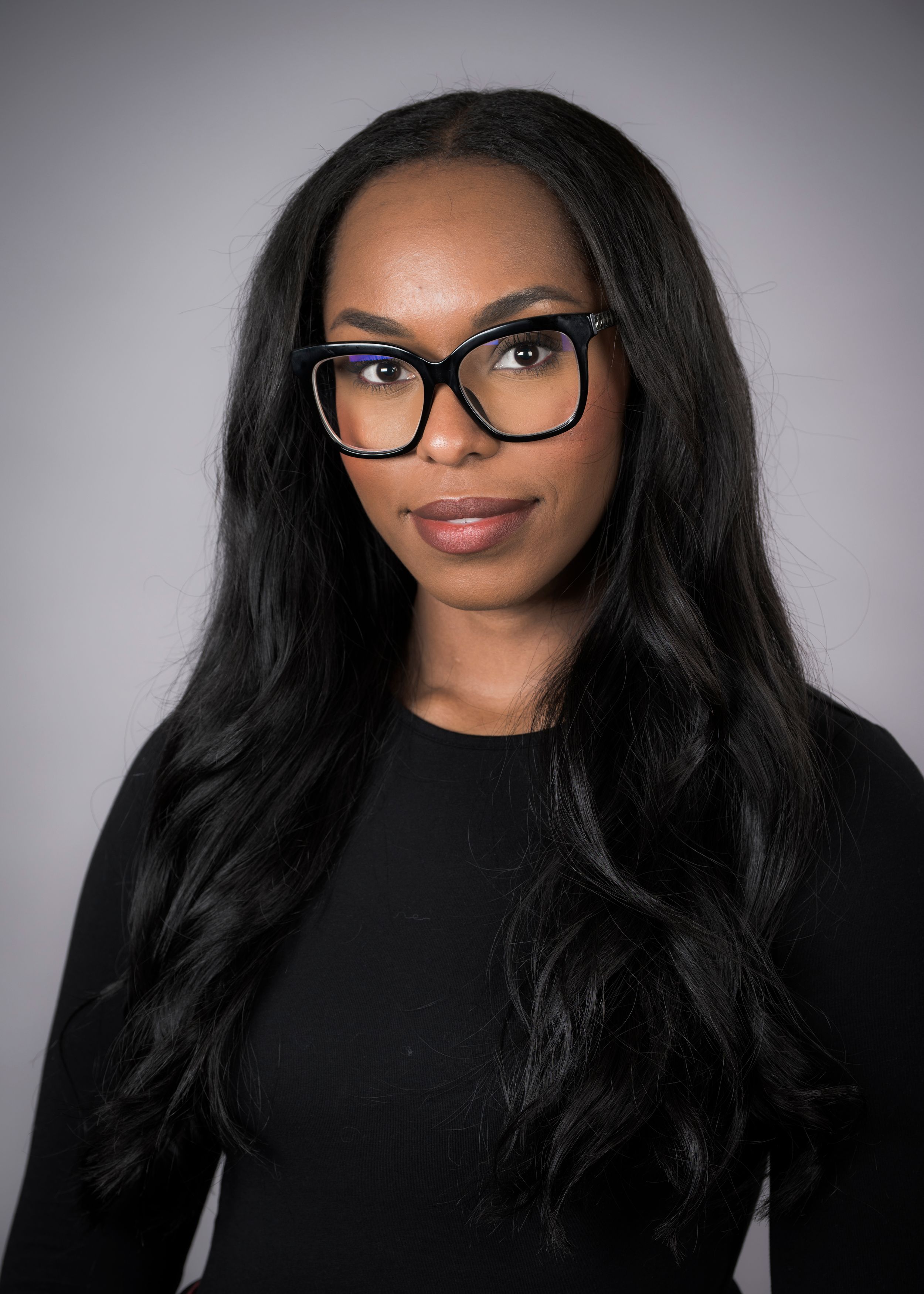 Professional portrait of someone wearing stylish black-framed glasses and a black top against a gray background.