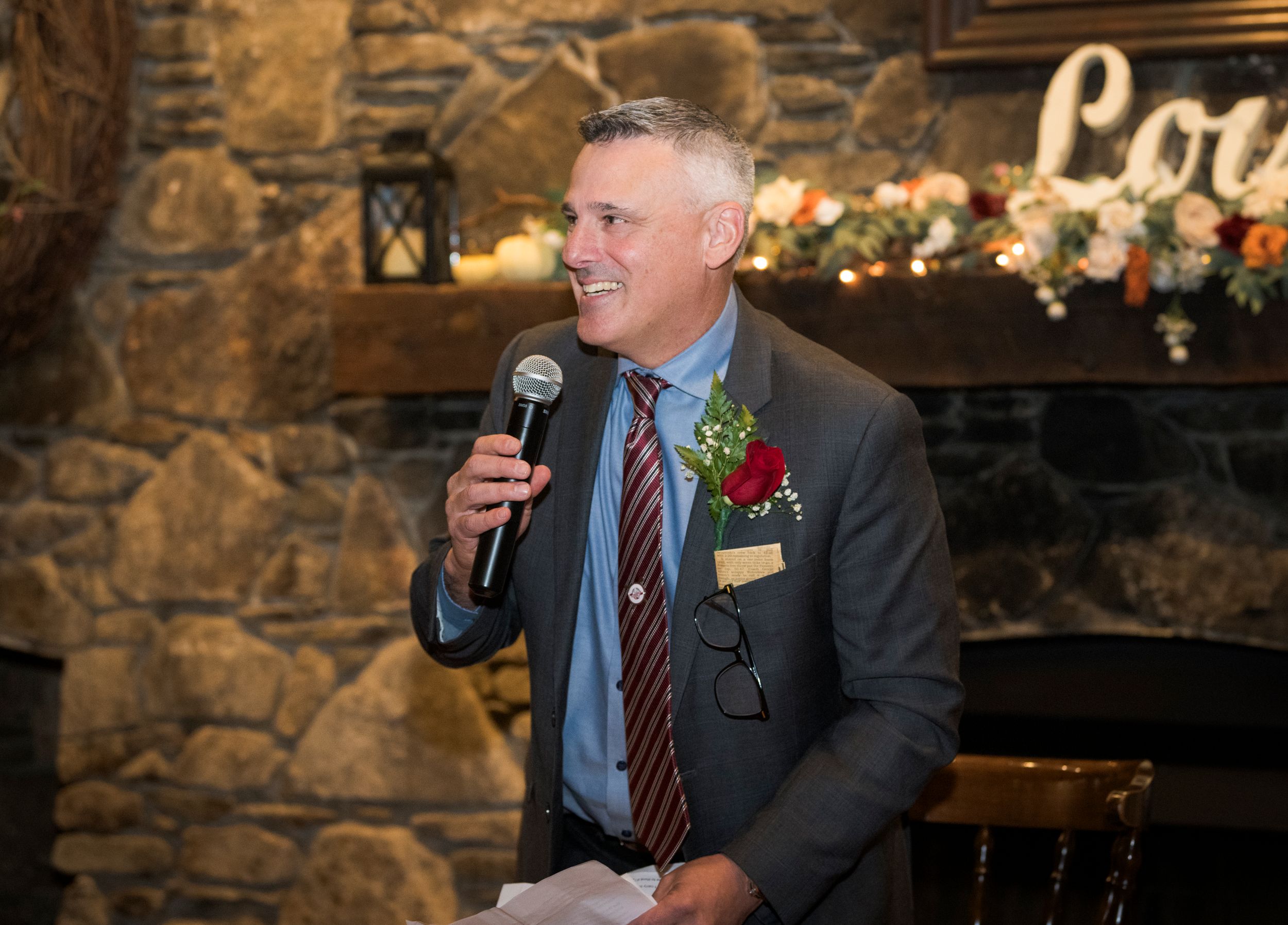 Someone in a gray suit and striped tie gives a speech with a microphone at an event with stone walls and twinkle lights.