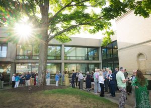 A panoramic view shows people gathered outside a building on a sunny day with trees casting dappled shade.