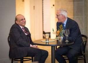 Two men in business attire sit at a small round table having an animated conversation over drinks.