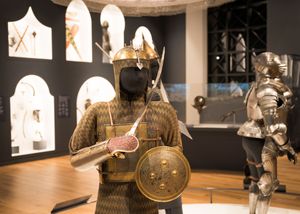 Medieval armor and weaponry displays arranged in a museum exhibition with dramatic lighting and dark backgrounds.