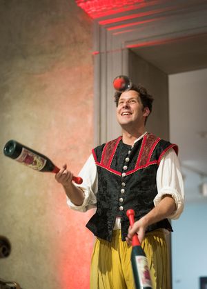 A performer in Renaissance costume juggles bottles while wearing a black vest and yellow pants on stage.