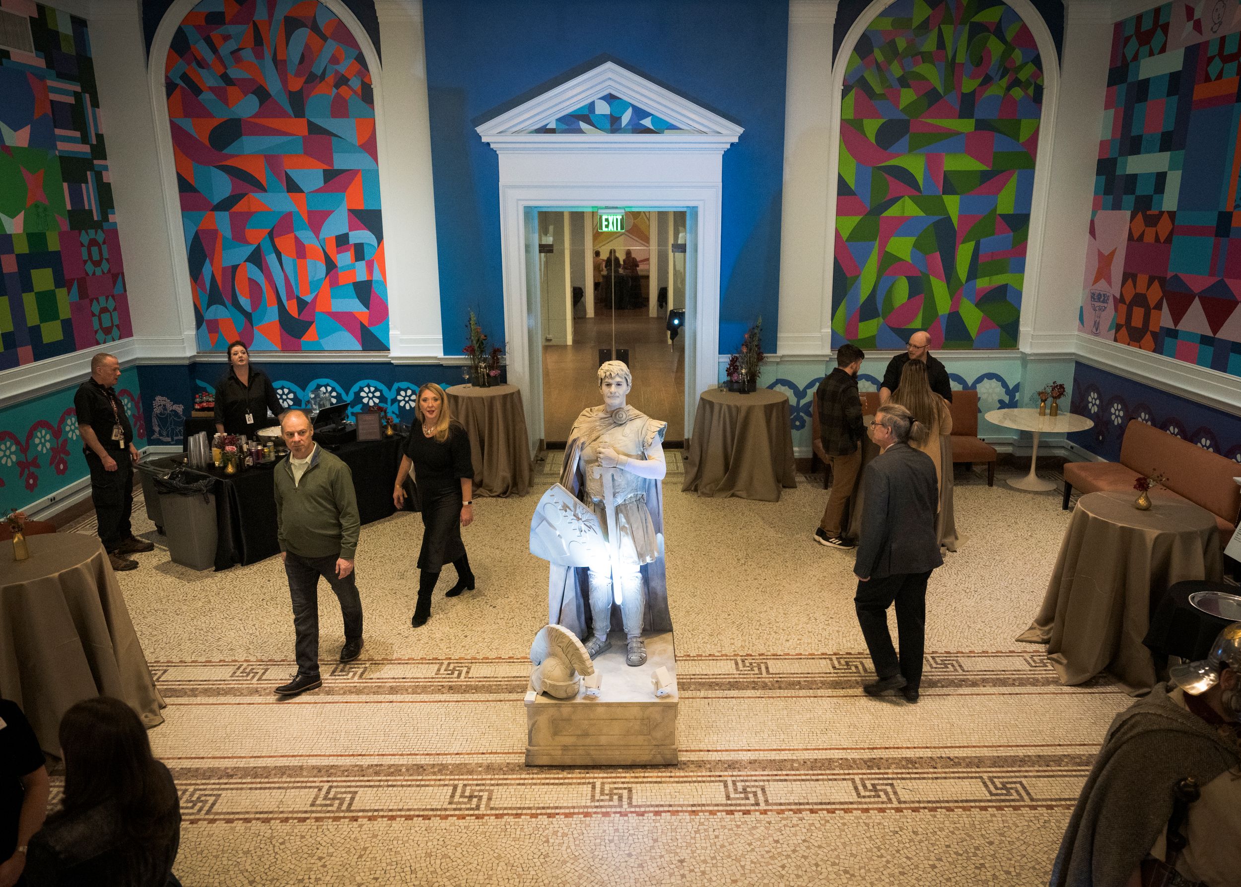 A performer in a flowing blue and white costume stands on stage while others gather around in a room with vibrant wall art.