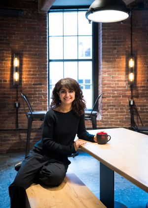 A person in black clothing sits and poses in a modern industrial office space with exposed brick walls and large windows.