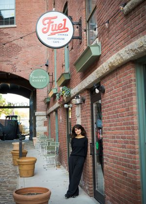 A figure in a long black dress poses against a rustic brick wall with vintage signage and potted plants in an urban setting.