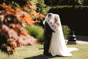 A couple in wedding attire shares a romantic moment in a garden setting with soft autumn foliage in the background.