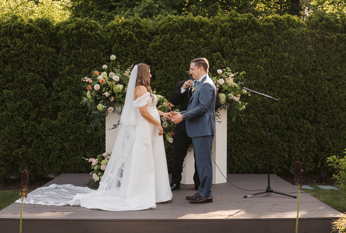 A wedding ceremony takes place in a lush garden setting with a decorative white arch and floral arrangements.