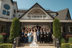 Large wedding party poses on grand staircase of elegant white mansion with ornate architecture and manicured landscaping.
