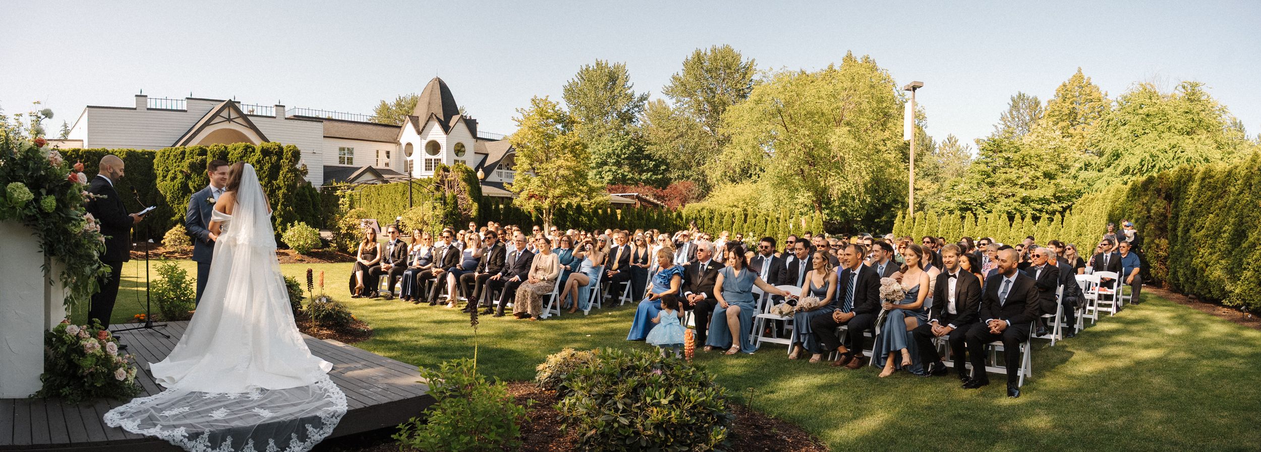 Panoramic view of an outdoor wedding venue with guests gathered under blue skies and green trees.