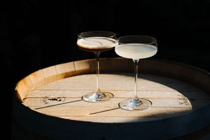 Two elegant cocktail glasses with different colored drinks sit on a rustic wooden serving board against a dark background.