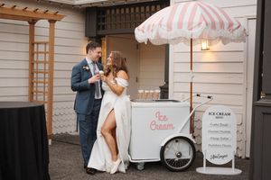 A vintage ice cream cart with pink and white striped umbrella stands next to a white building.
