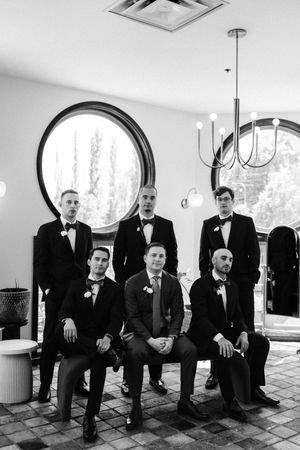 A formal group portrait of groomsmen in black tuxedos seated and standing in front of an arched window.