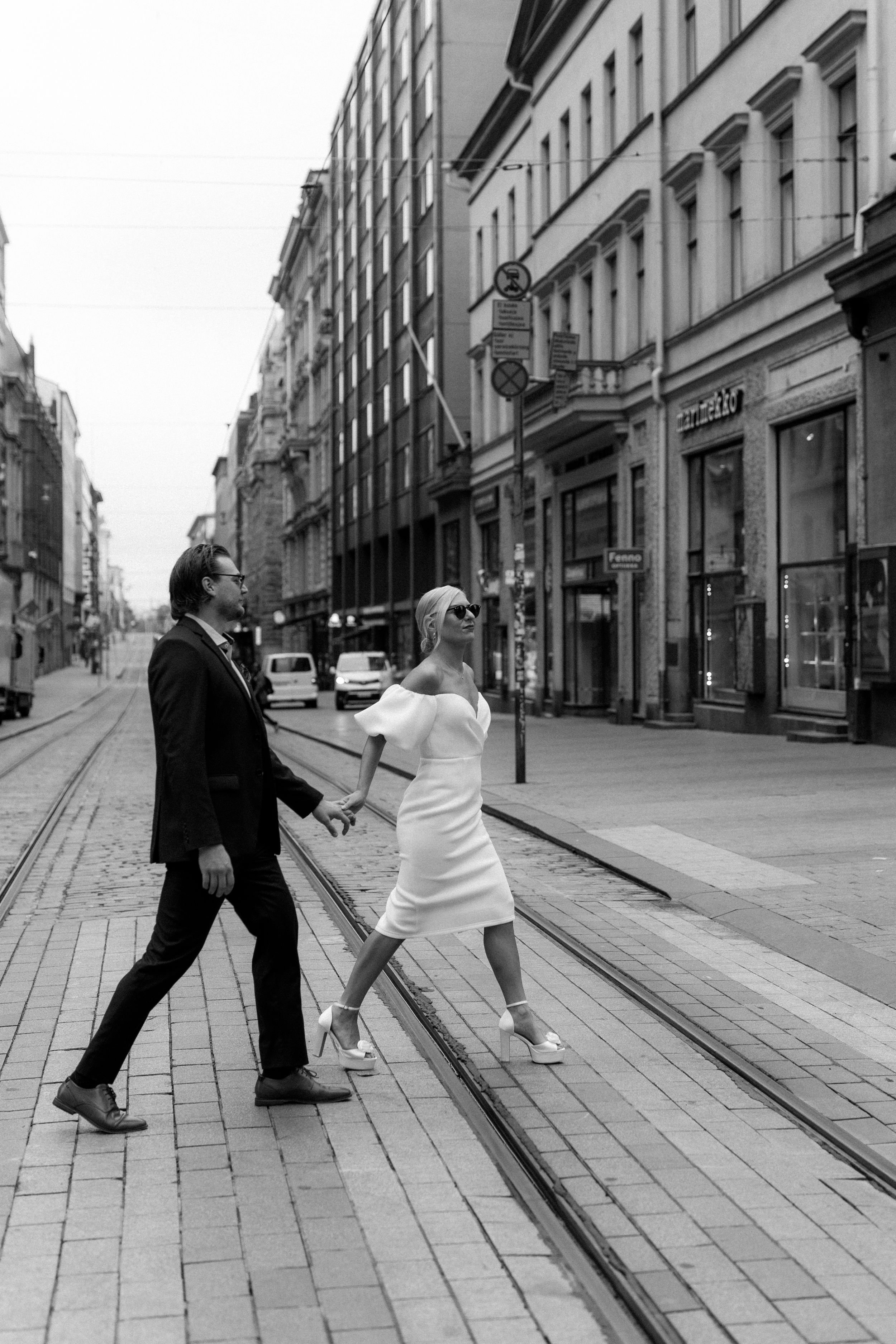 Mustavalkoinen hääpotretti jossa tyylikkäästi pukeutunut hääpari kävelee käsi kädessä suojatien yli Aleksanterinkadulla Helsingissä