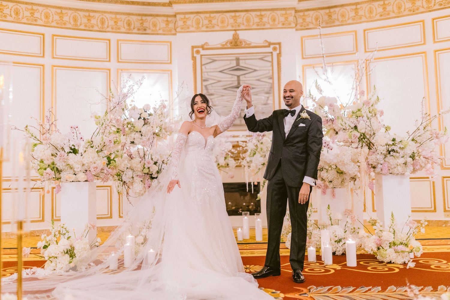 Elegant wedding ceremony in a luxurious white and gold ballroom decorated with white floral arrangements and chandeliers.
