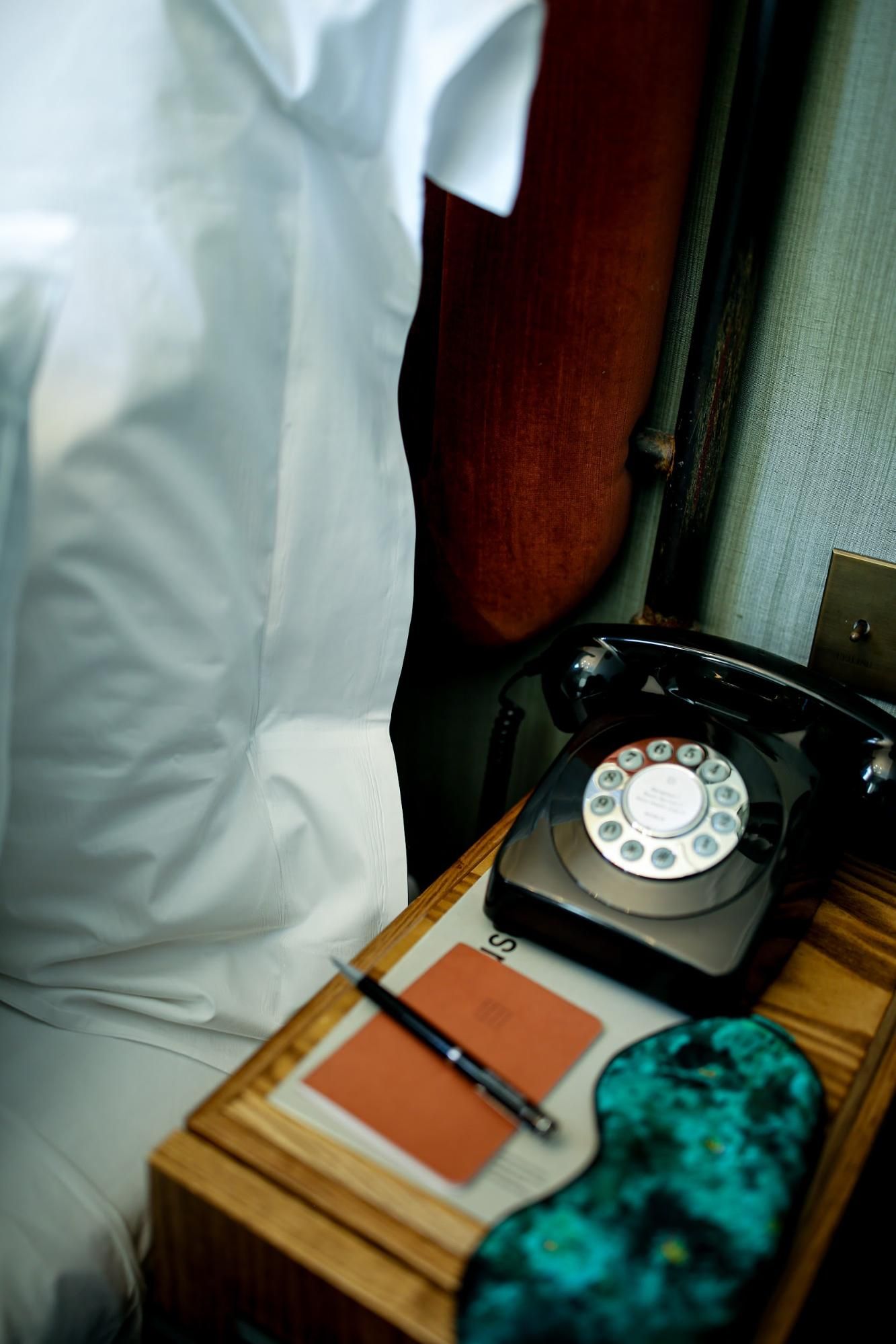 A vintage rotary phone sits on a wooden desk with white curtains and teal decor in the background.