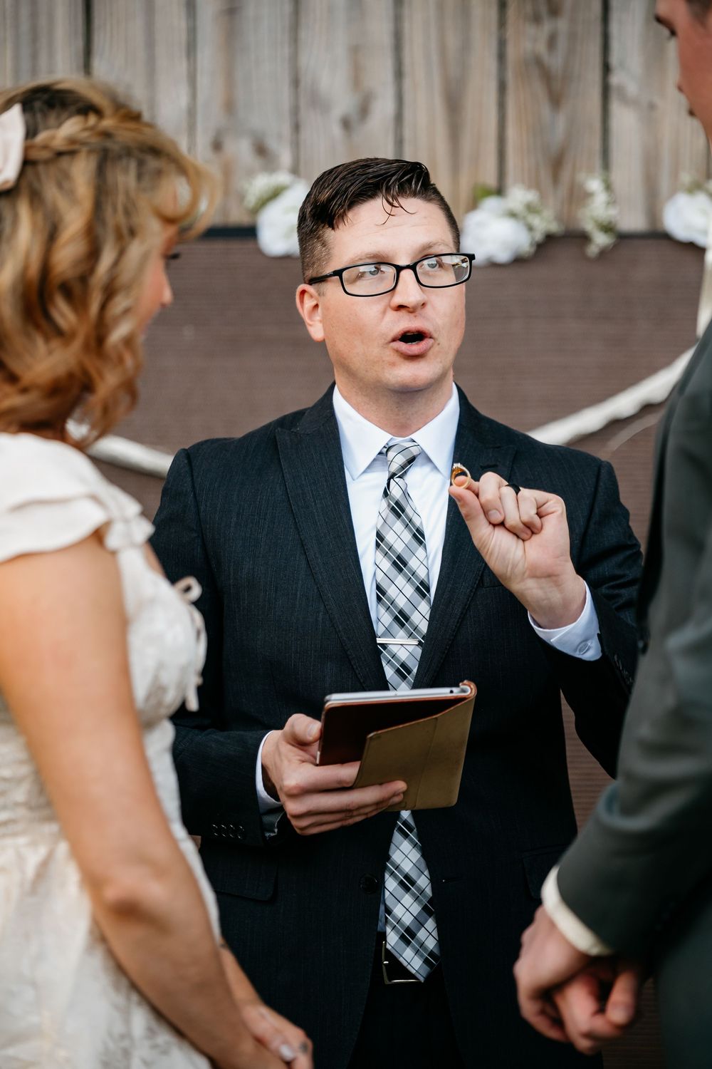 A young boy in a suit and tie gestures excitedly while holding a clipboard during a wedding ceremony.