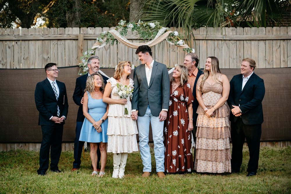 Wedding party laughs together while standing in a line against a wooden fence during an outdoor celebration.