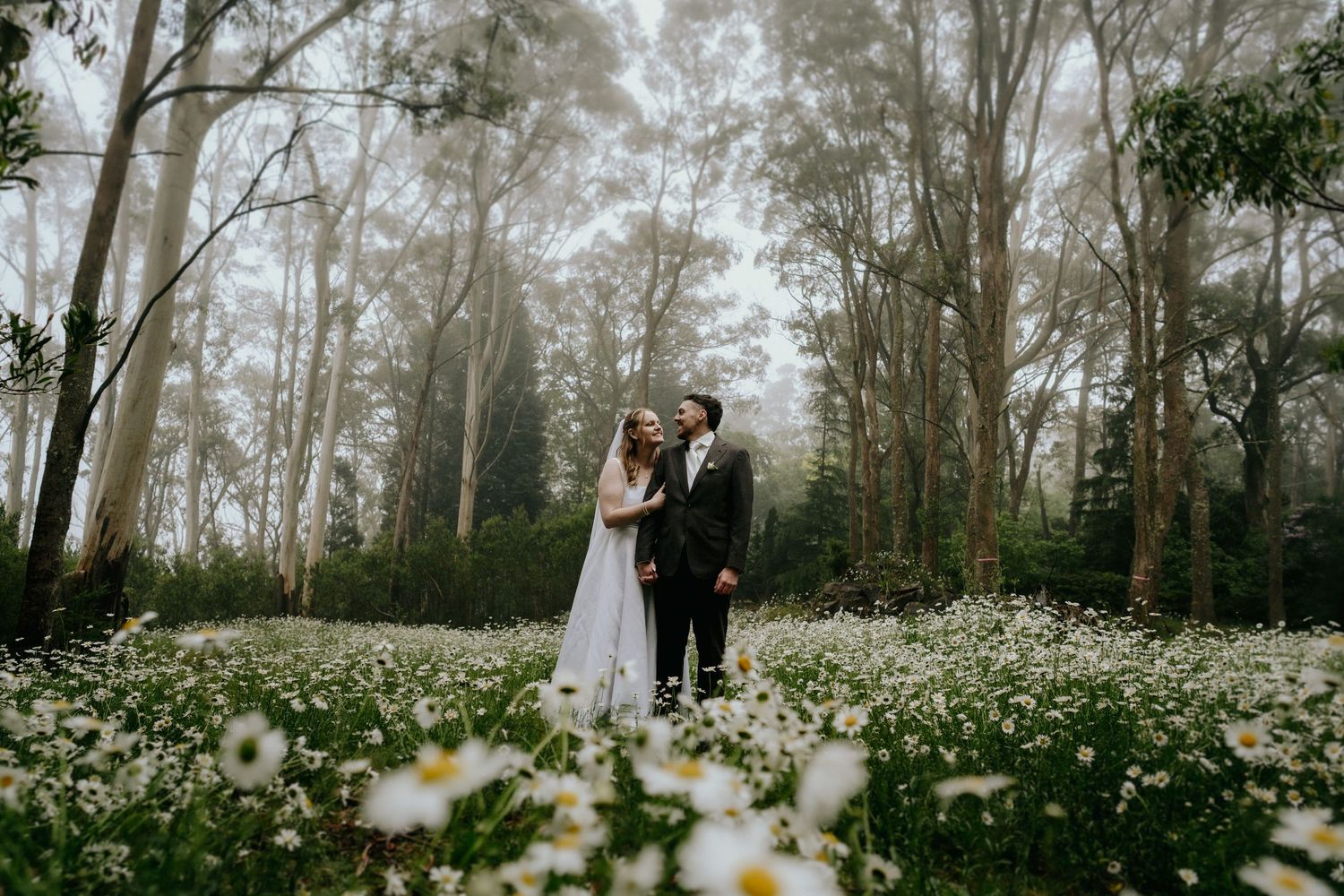 Wedding couple stands among blooming wildflowers in ethereal forest setting.