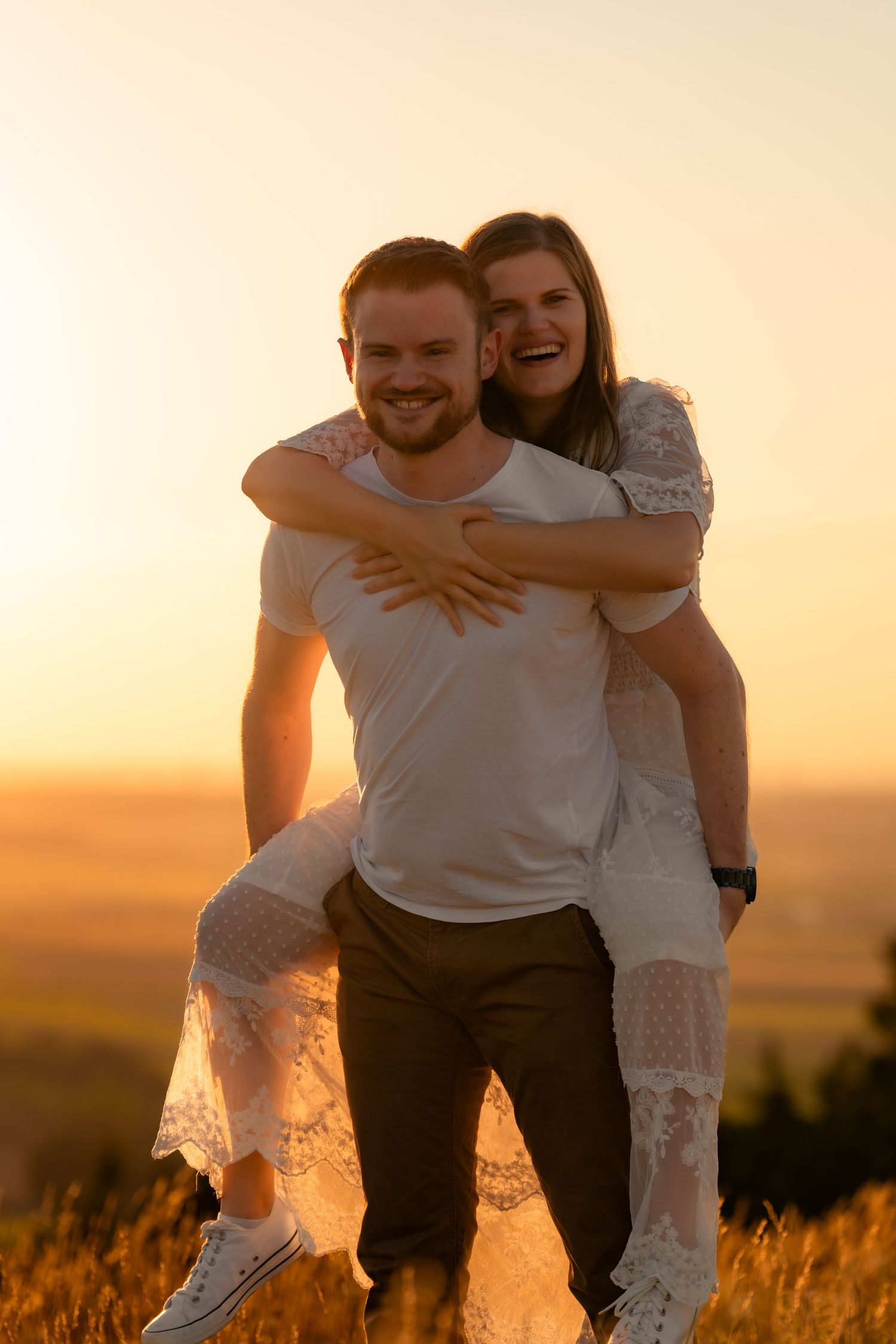 A couple shares a joyful piggyback ride moment in a golden field during sunset.