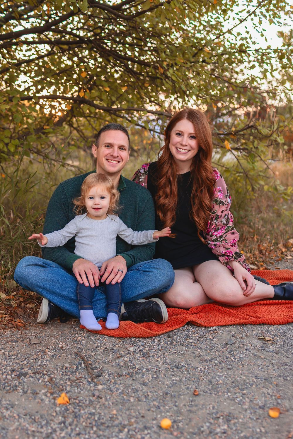 Family of 3 mom, dad and toddler girl sitting together on orange blanket during autumn photoshoot with fall foliage in background.