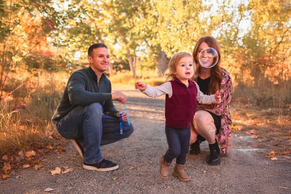 Family of 3 mom, dad and toddler daughter sat on a dirt path playing with bubbles surrounded by golden autumn foliage at sunset.