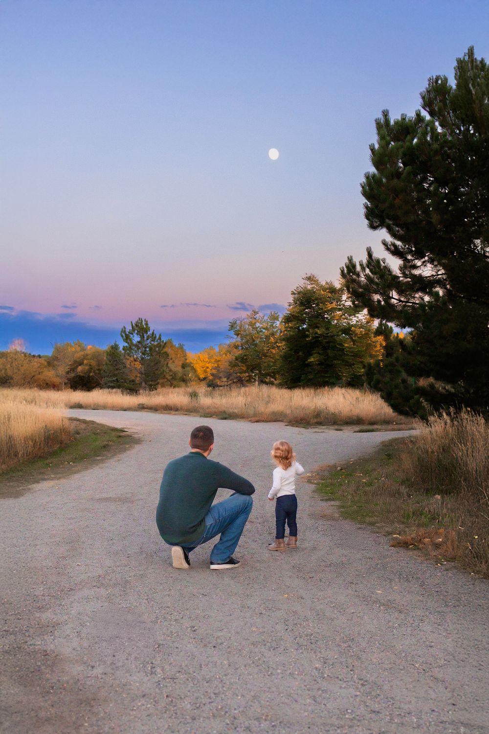 Father and toddler daughter sit on a dirt path at sunset while observing the full moon rising in the autumn sky.