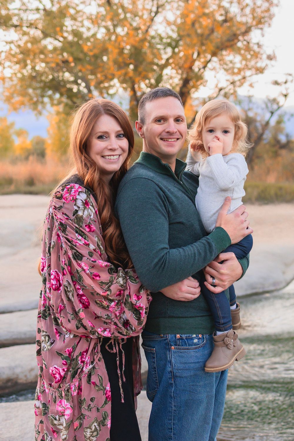 A family poses together outdoors on a dirt path with autumn trees and golden foliage in the background.  Mom, Dad and toddler daughter