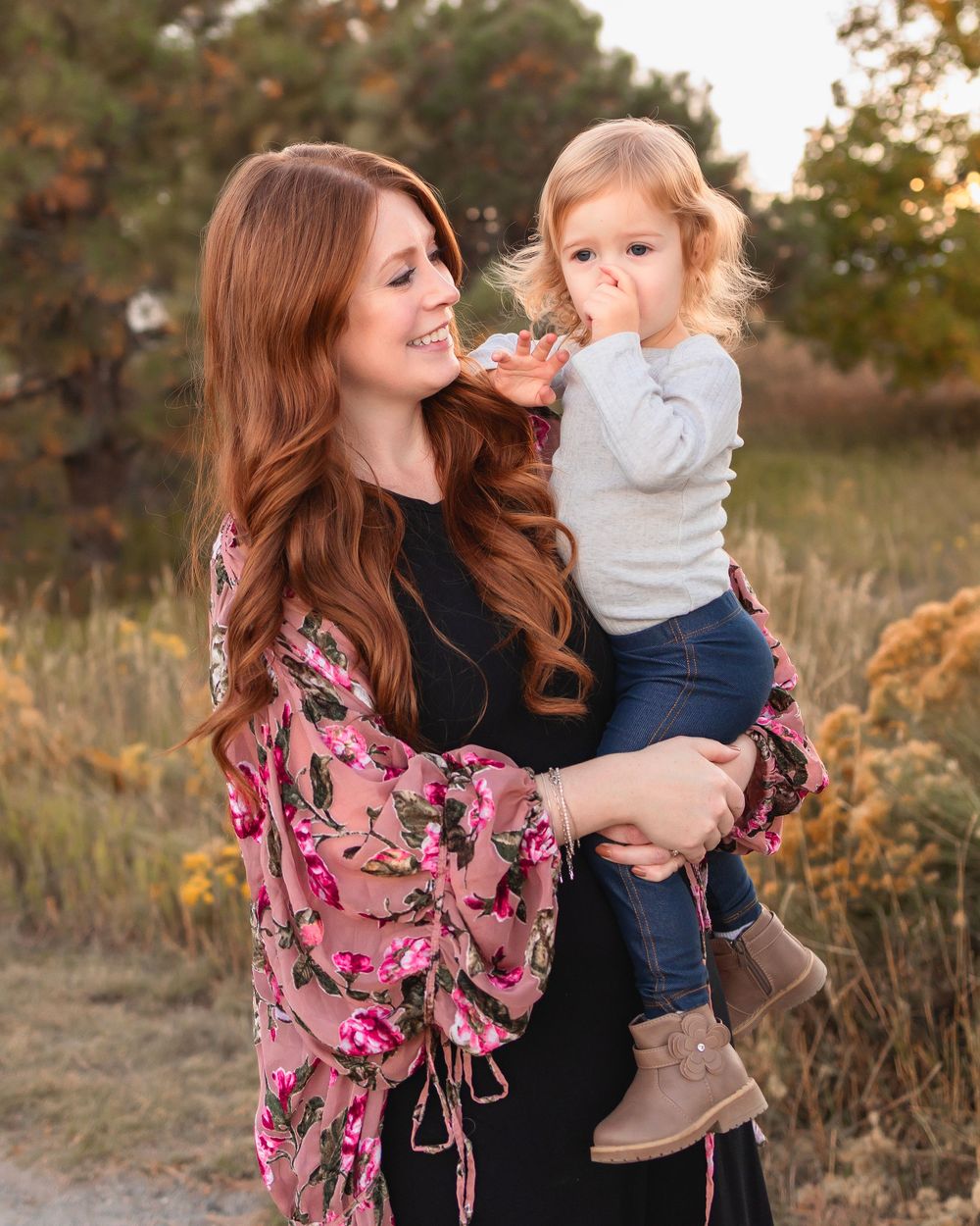 A woman in a floral pink kimono holds a small child against a sunset backdrop of tall grass and trees.