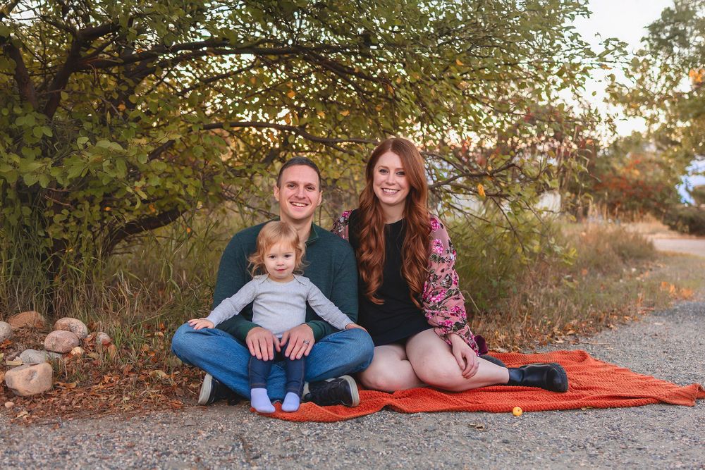 A family of three, mom, dad and toddler daughter sits together on a red blanket during an outdoor autumn photo session in a natural setting.