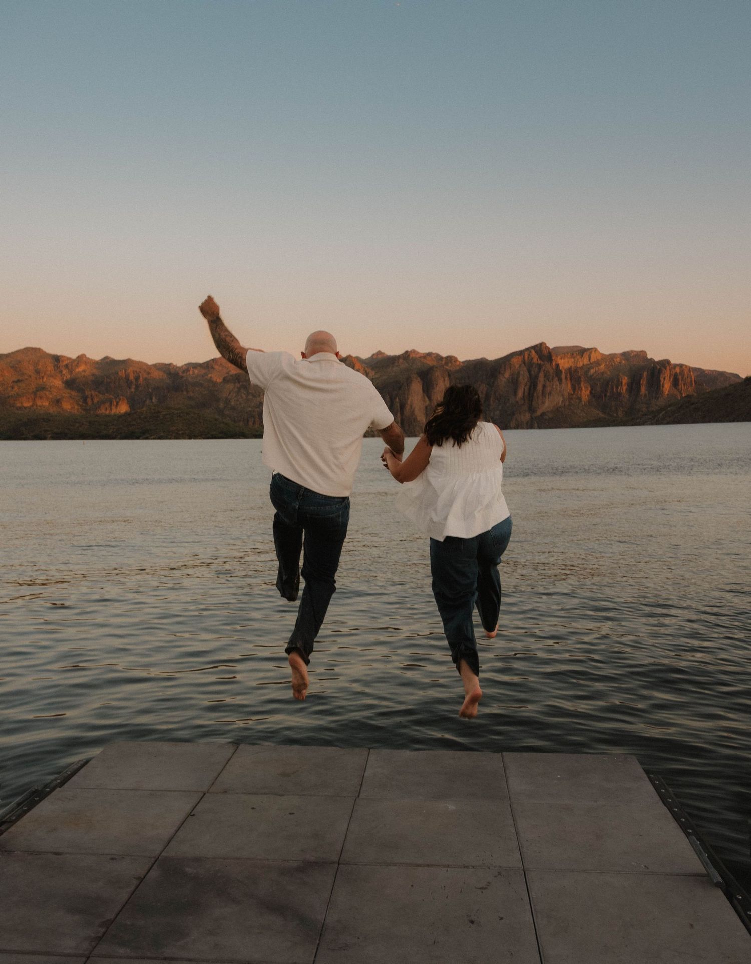 Couple jumping joyfully on a dock at sunset with mountain lake views behind them.