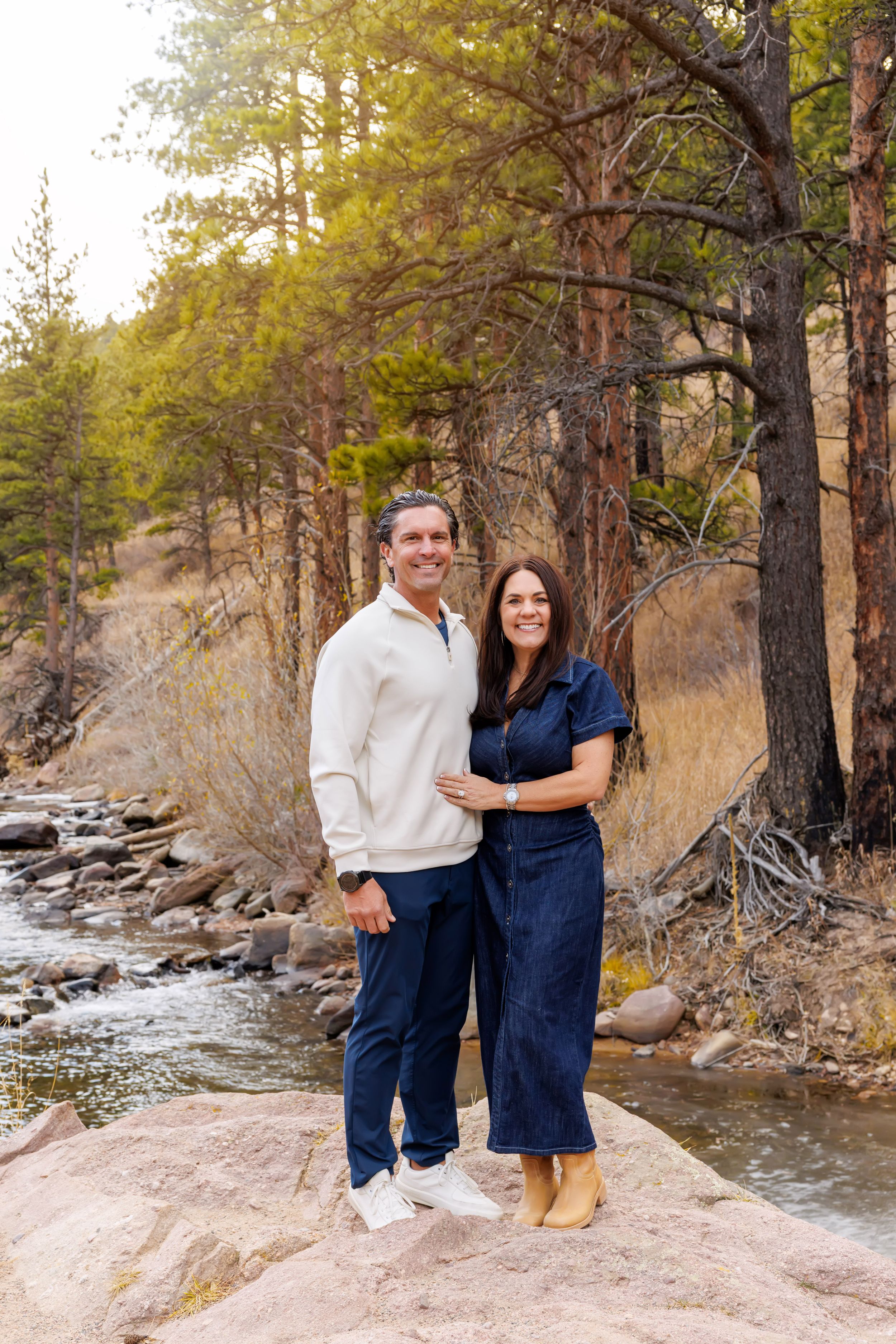 A couple in coordinating navy and white outfits poses together by a scenic mountain stream on a sunny autumn day.