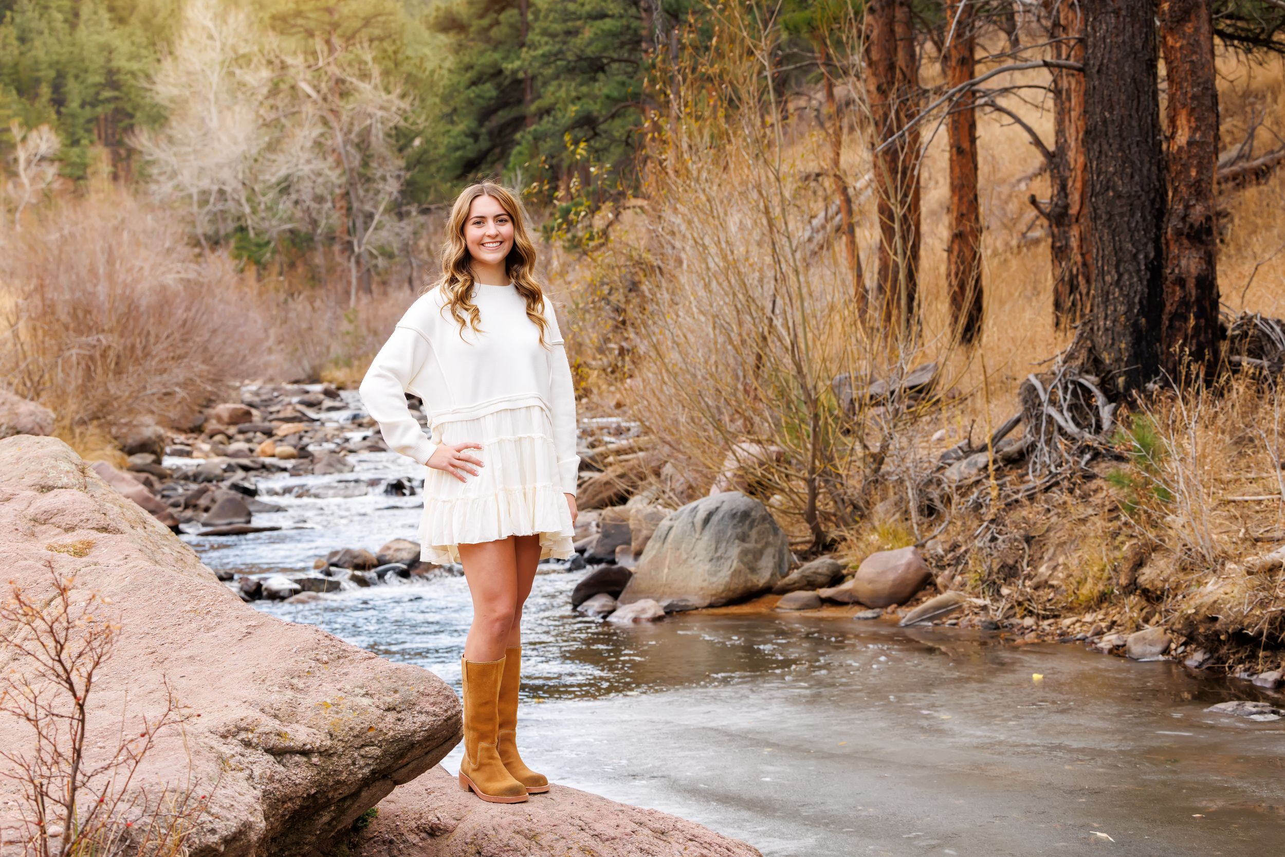 Person in a white dress stands on a rock by a scenic mountain stream during autumn.