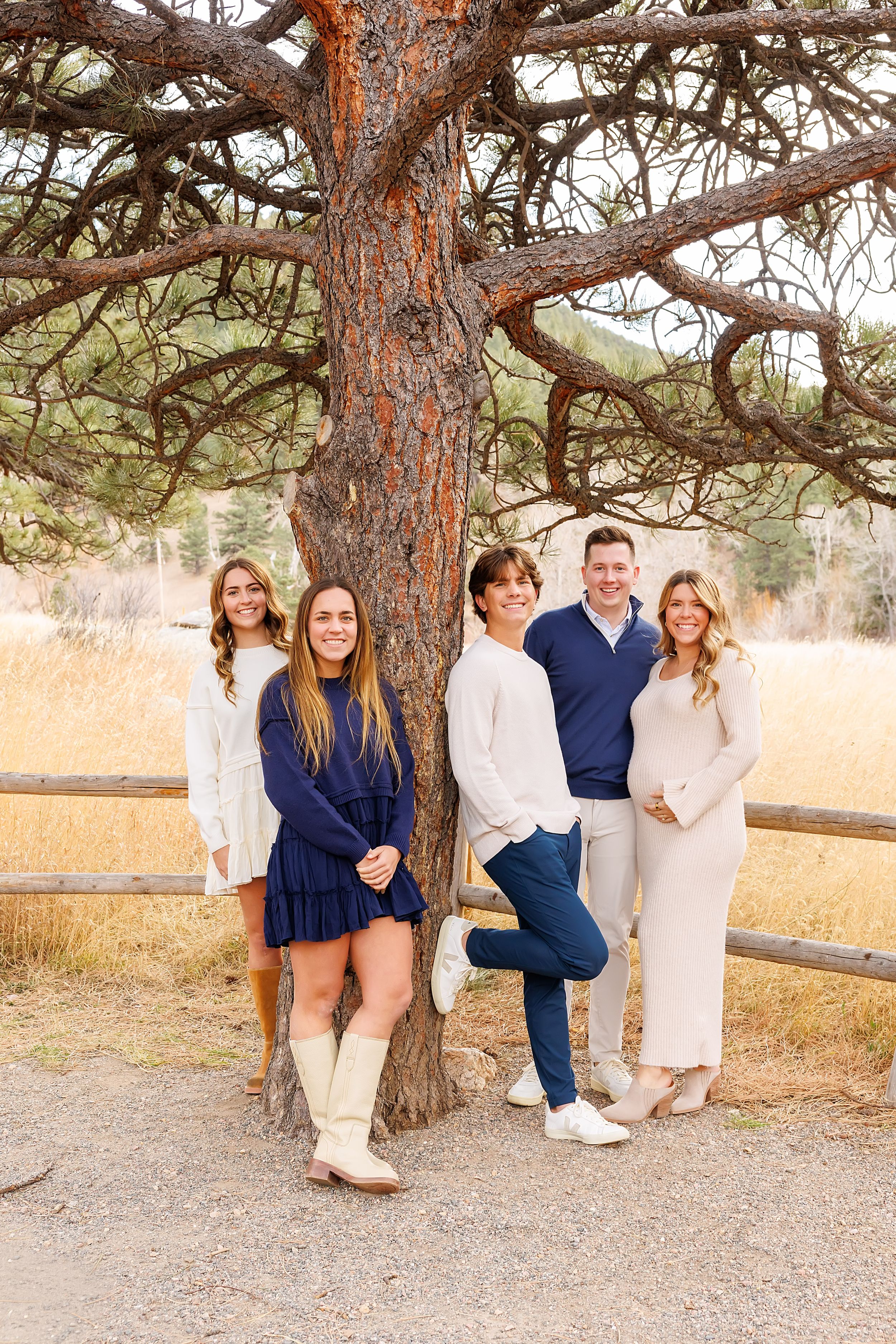 A group poses together in casual attire against a large pine tree with a rustic wooden fence and golden field behind them.