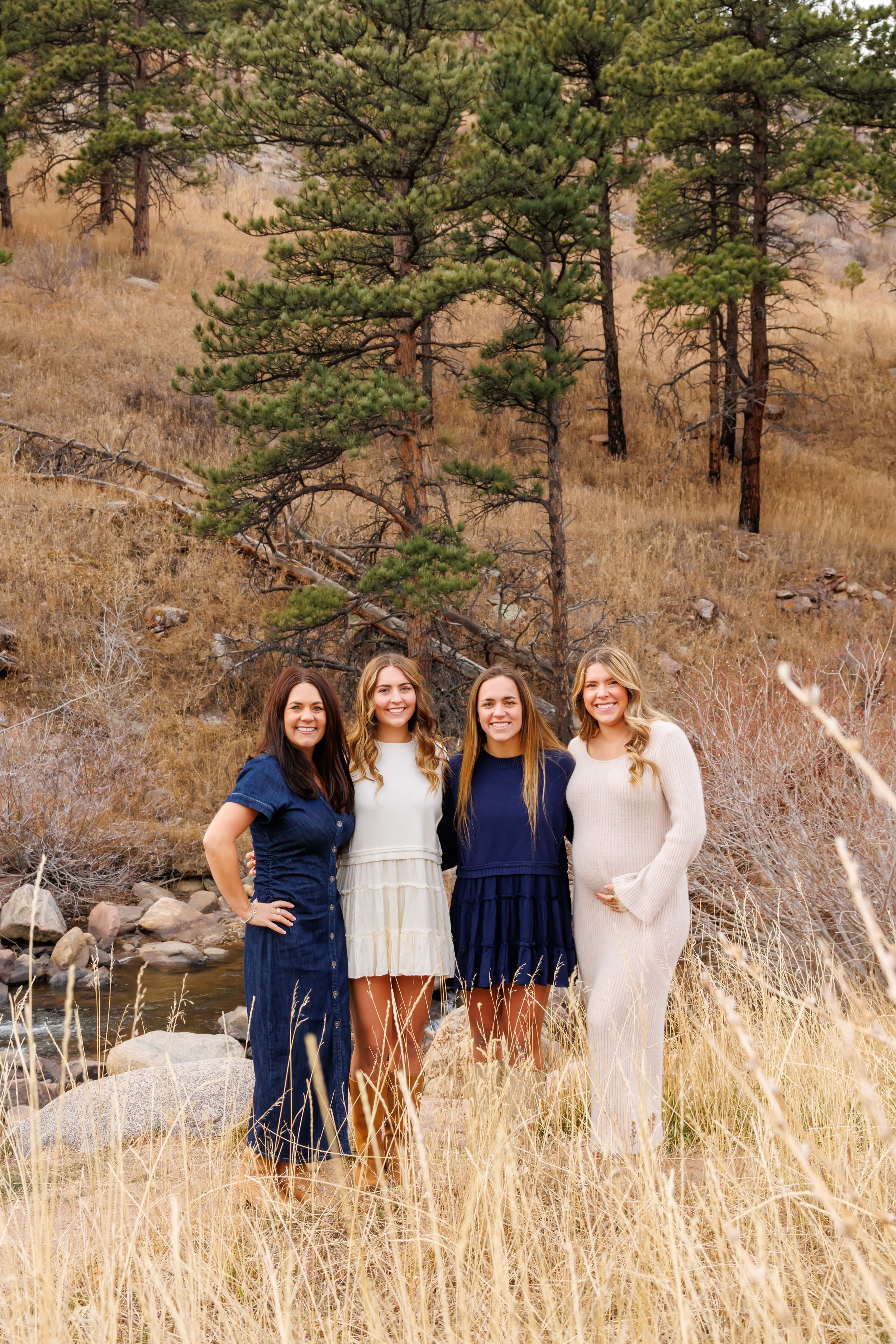 Four women in coordinating blue and white outfits pose together in a scenic mountain setting with pine trees and tall grass.