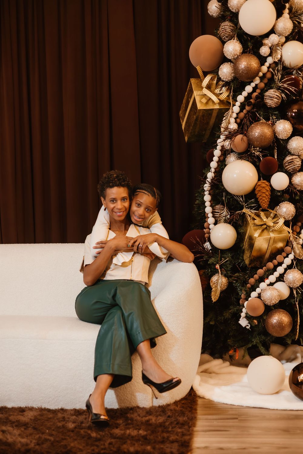 Two people share a tender moment on a white couch next to an elegant Christmas tree decorated in gold and white ornaments.