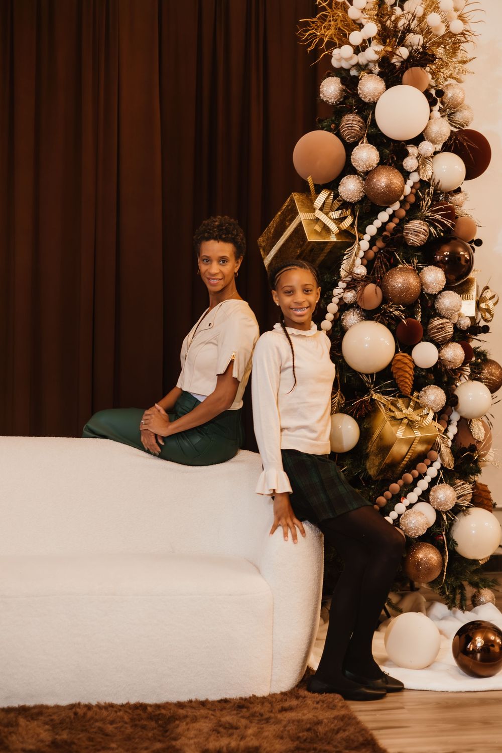 Two people pose back-to-back on a white sofa beside a Christmas tree adorned with gold and white decorations.