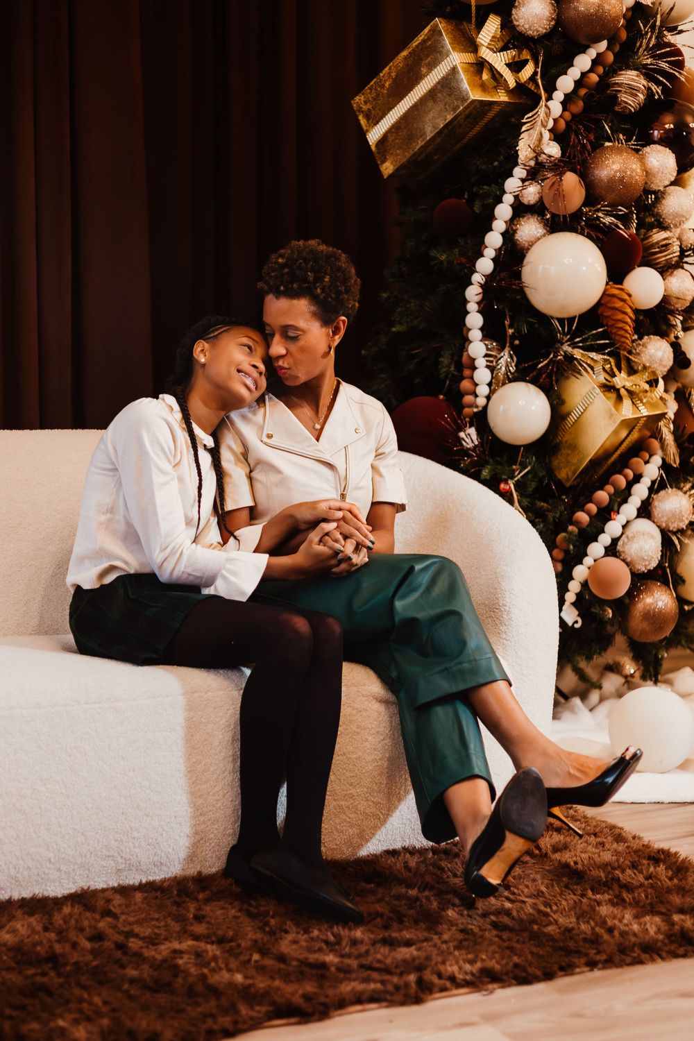A couple sits close together on a white couch next to an elegant Christmas tree decorated with gold and white ornaments.