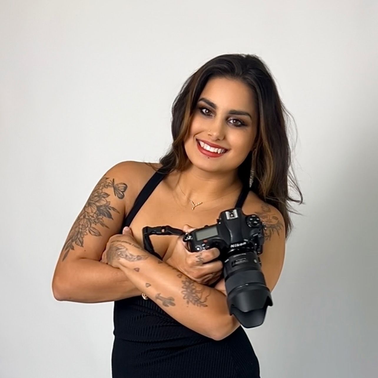 Professional photographer in black tank top holding DSLR camera and posing with tattoos visible in studio setting.