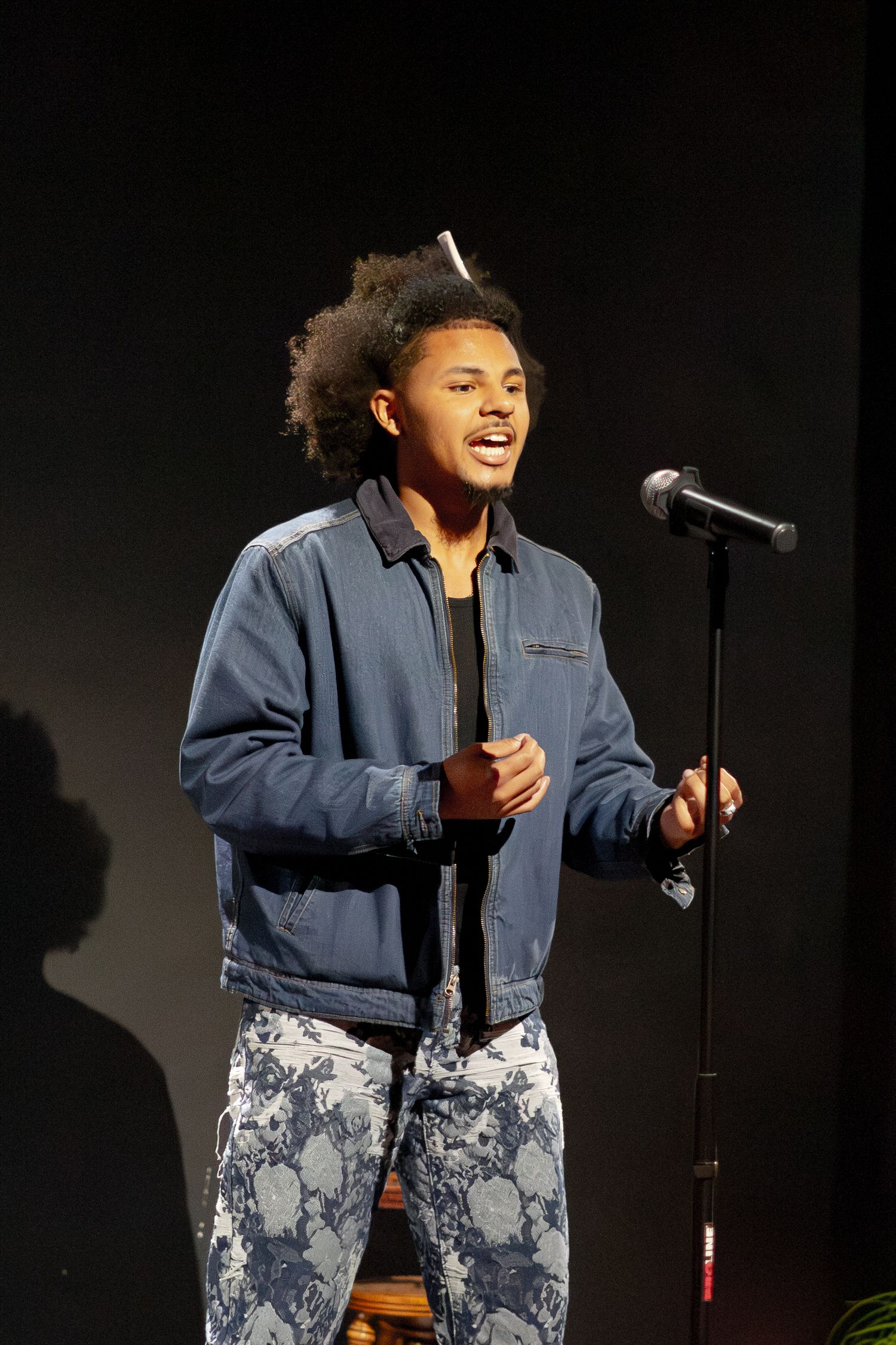 A performer in denim jacket and patterned pants stands on stage with a microphone.