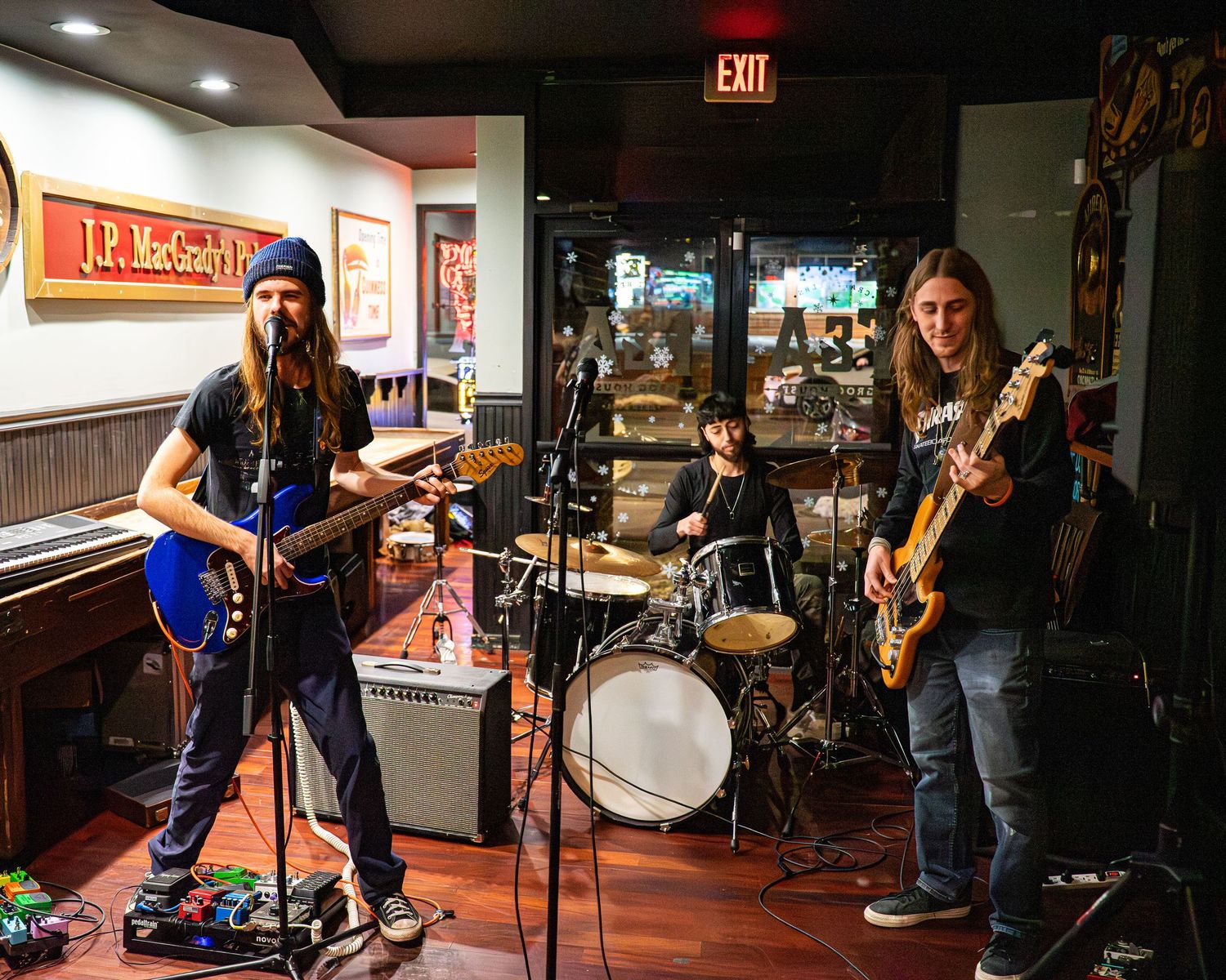 Wide shot of band performing on stage in a bar with exit sign visible.